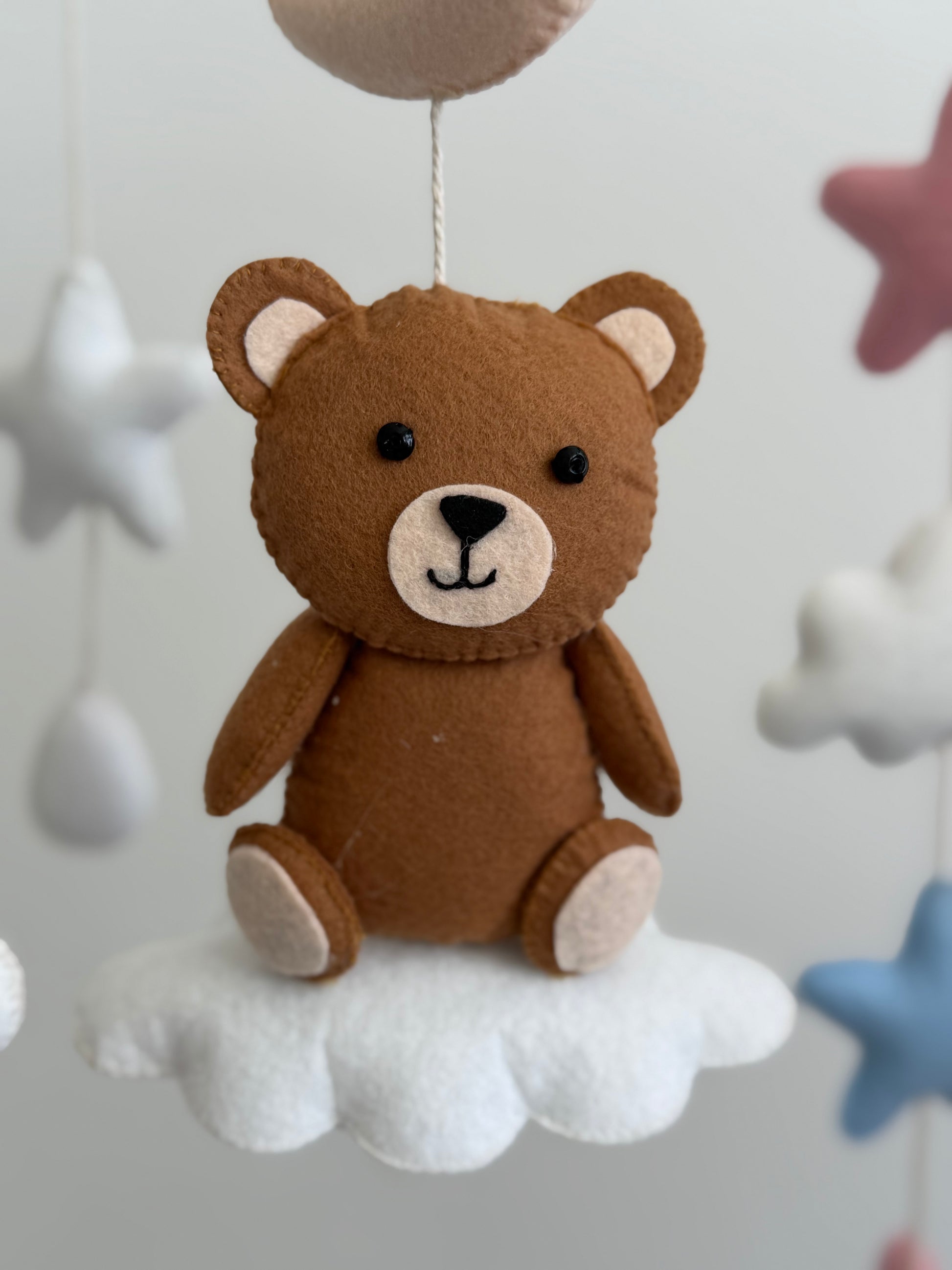 Brown teddy bear toy hanging from a cloud against a light gray background with star decorations.