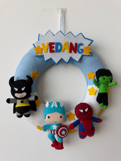 Decorative wreath with superhero-themed plush toys and 'VEDANG' sign on a white background