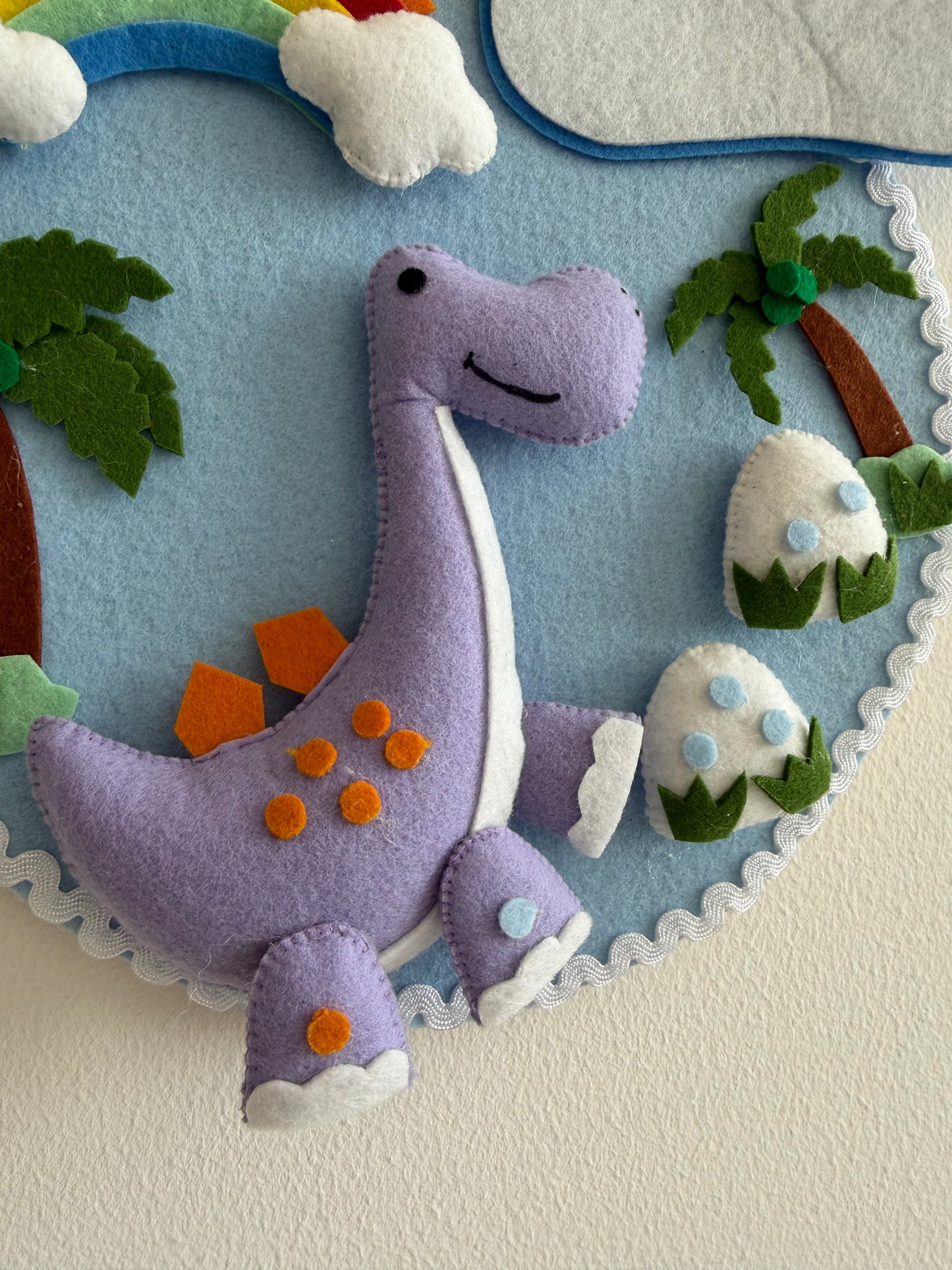 Felt dinosaur toy with a colorful background featuring trees and eggs.