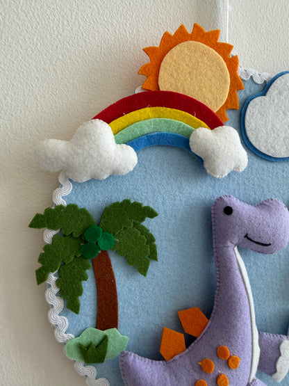 Felt wall hanging with a dinosaur, rainbow, sun, clouds, and palm trees on a light blue background.